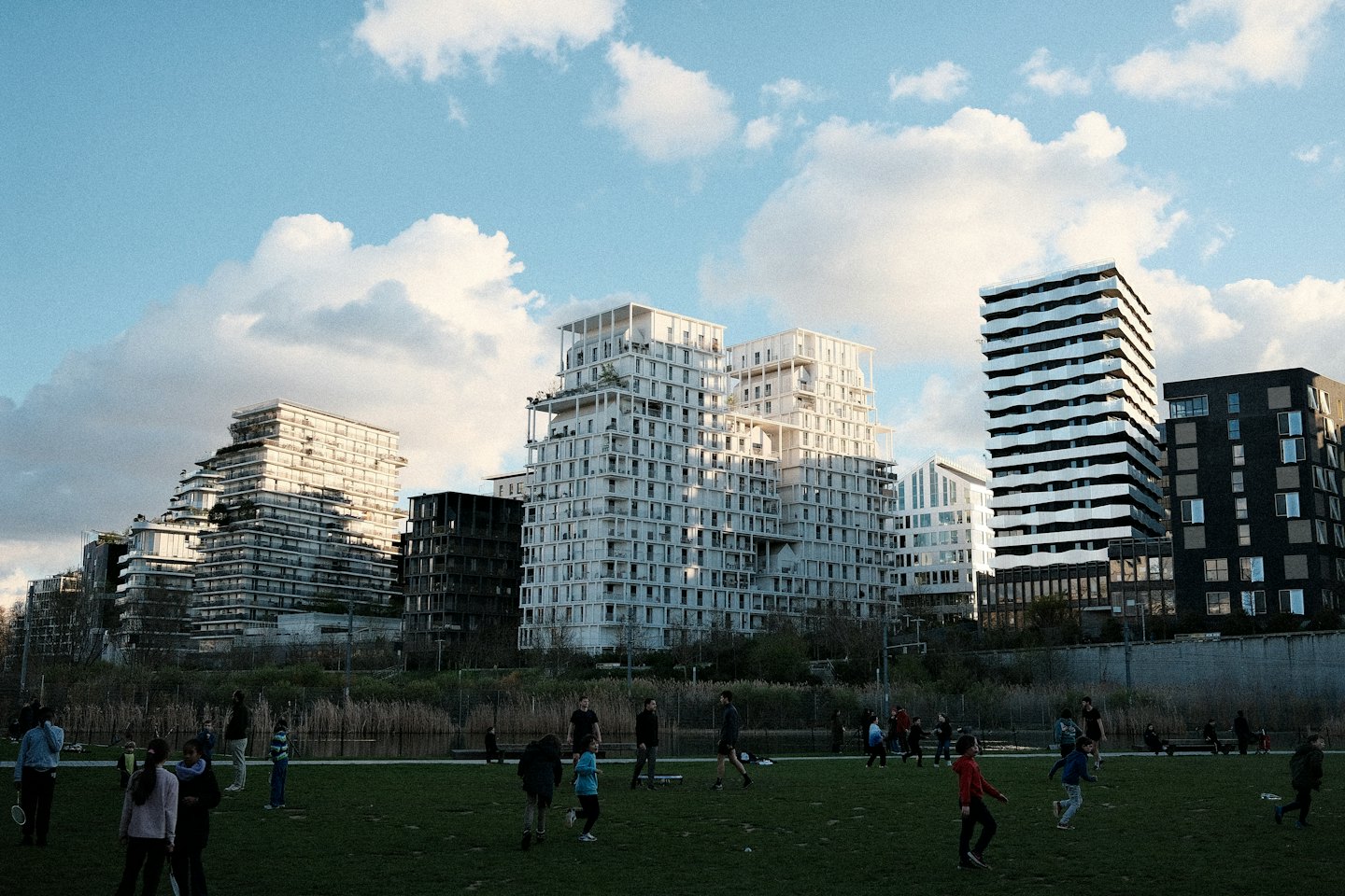 People playing badminton on a sunny park field with city buildings behind them.